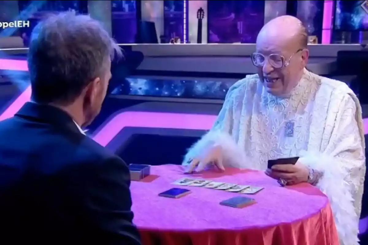 Rappel con gafas y túnica blanca está sentado en una mesa redonda cubierta con un mantel rojo, frente a Pablo Motos, mientras sostiene cartas y parece estar realizando una lectura de tarot en un ambiente iluminado con luces de neón. Rappel con gafas y túnica blanca está sentado en una mesa redonda cubierta con un mantel rojo, frente a Pablo Motos, mientras sostiene cartas y parece estar realizando una lectura de tarot en un ambiente iluminado con luces de neón.