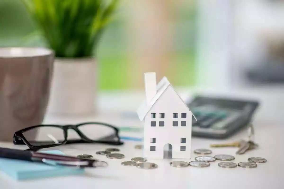 Imagen de una maqueta de una casa en miniatura con varias monedas y unas gafas y una calculadora alrededor Imagen de una maqueta de una casa en miniatura con varias monedas y unas gafas y una calculadora alrededor