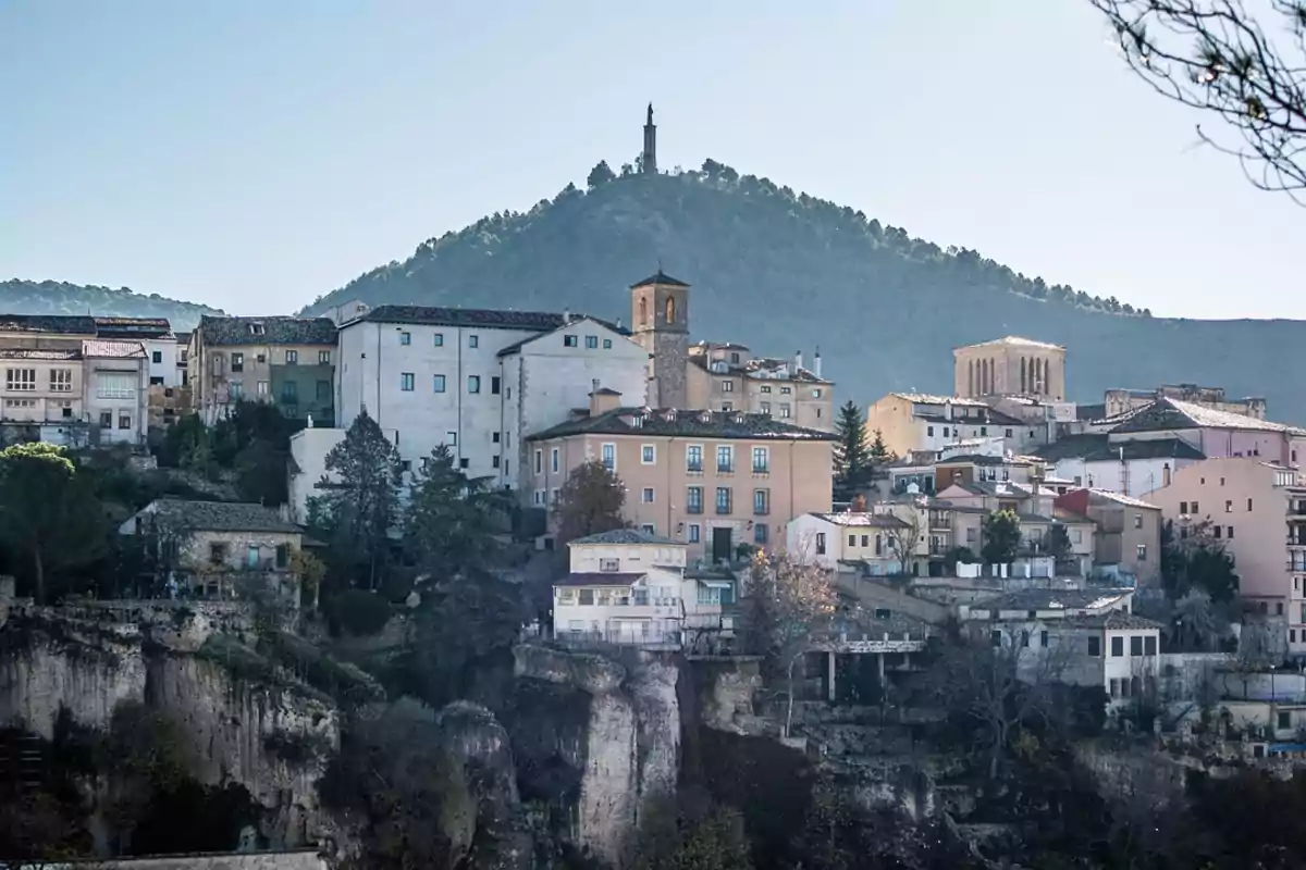 Vista de un pueblo con casas antiguas construidas sobre un acantilado y una colina boscosa al fondo coronada por una estatua