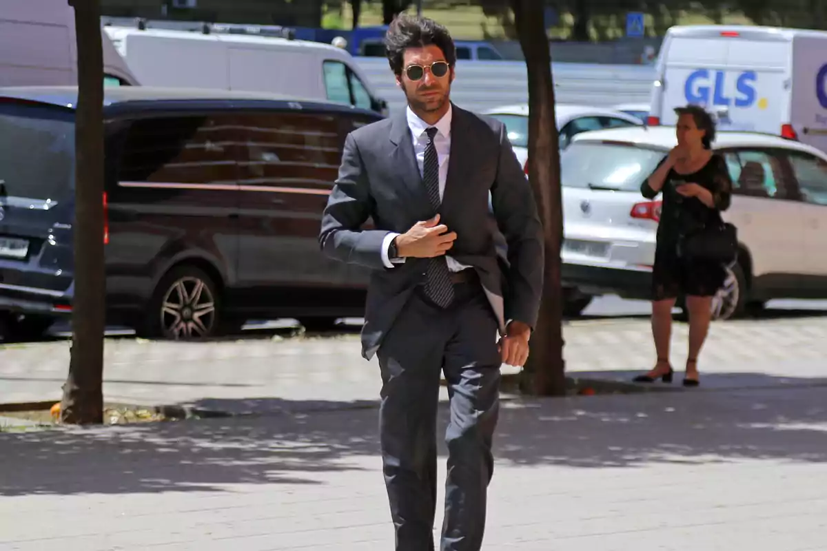 Cayetano Rivera con traje y gafas de sol camina por la acera en un entorno urbano con vehículos estacionados al fondo. Cayetano Rivera con traje y gafas de sol camina por la acera en un entorno urbano con vehículos estacionados al fondo.