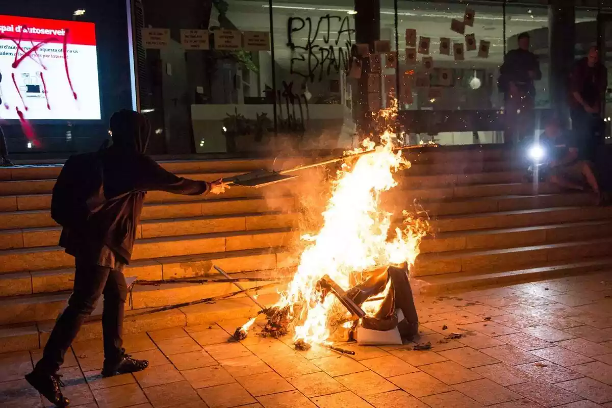 Plano general de un manifestante independentista vestido de negro y encapuchado lanzando un objeto a una hoguera situada delante de un edificio que tiene un grafiti que pone 'Puta Espanya' y una estelada