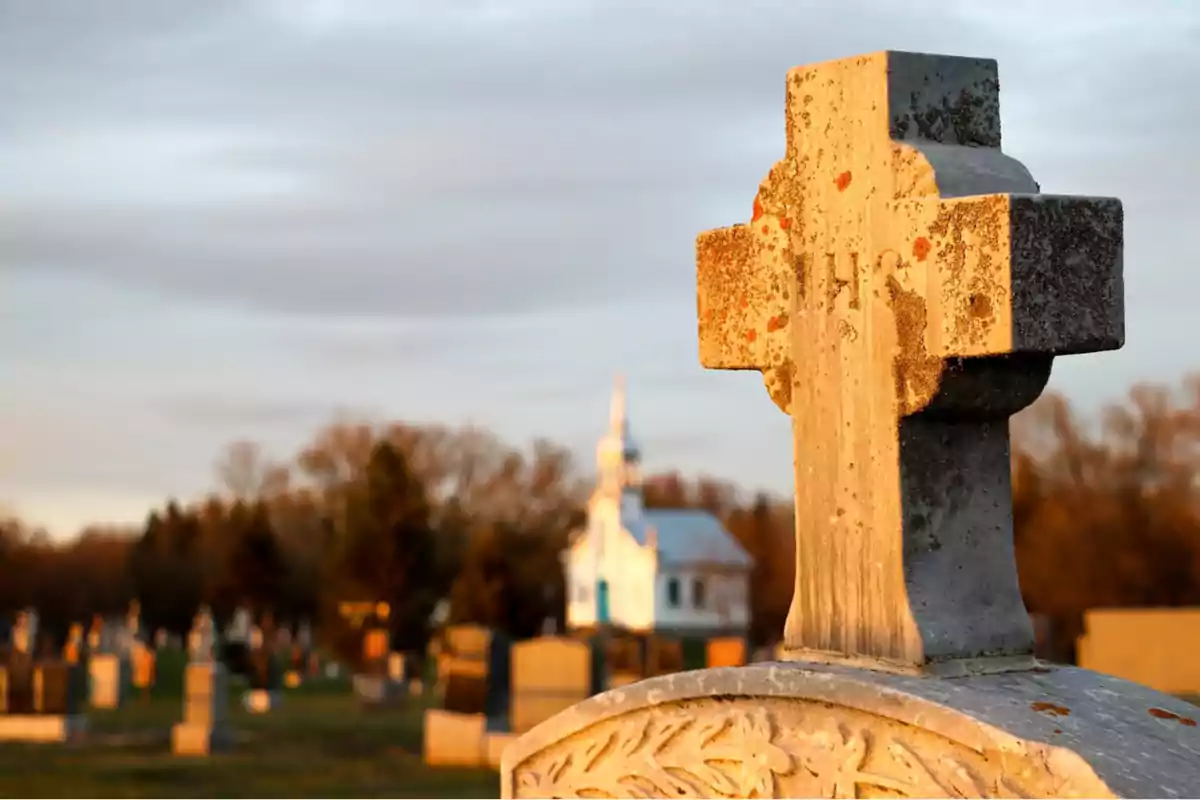 Una cruz de piedra en un cementerio con una iglesia al fondo al atardecer.