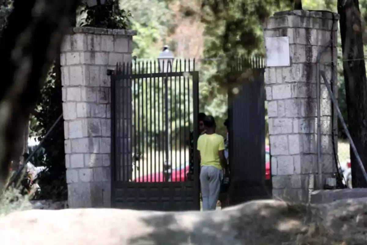 Una persona con camiseta amarilla y pantalones grises está de pie frente a una puerta de hierro entre dos pilares de piedra en un entorno arbolado.