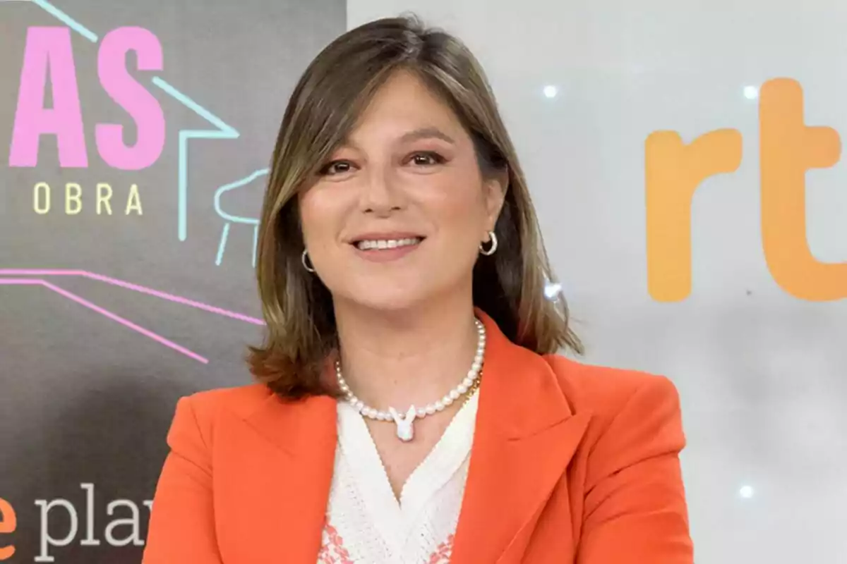 Captura de Chabeli Iglesias sonriente con cabello castaño y lacio, vestida con un blazer naranja y un collar de perlas, posa frente a un fondo con logotipos y texto colorido en la rueda de prensa de su programa Los Iglesias: Hermano a la obra