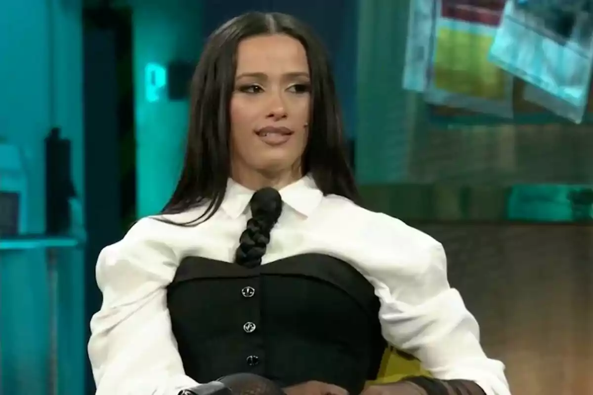 Chanel con cabello largo y oscuro, vestida con una camisa blanca y un corsé negro, está sentada en un entorno interior iluminado en 'La Revuelta'.