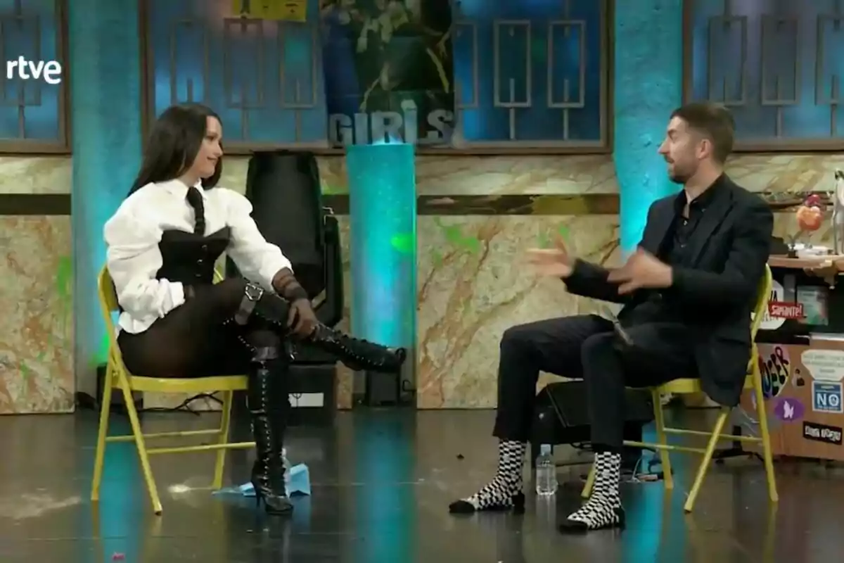 Chanel y David Broncano están sentadas en sillas amarillas en un set de televisión, una de ellas lleva un atuendo blanco y negro con botas altas, mientras que la otra viste un traje oscuro y calcetines a cuadros en 'La Revuelta'. Chanel y David Broncano están sentadas en sillas amarillas en un set de televisión, una de ellas lleva un atuendo blanco y negro con botas altas, mientras que la otra viste un traje oscuro y calcetines a cuadros en 'La Revuelta'.