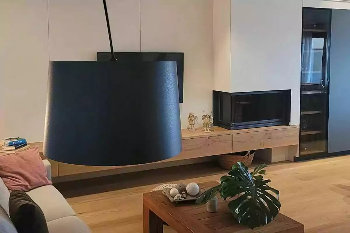 Una sala de estar moderna con una lámpara colgante negra, un sofá con cojines, una mesa de centro de madera con decoraciones, una planta, una televisión montada en la pared, una chimenea y una estantería. Una sala de estar moderna con una lámpara colgante negra, un sofá con cojines, una mesa de centro de madera con decoraciones, una planta, una televisión montada en la pared, una chimenea y una estantería.