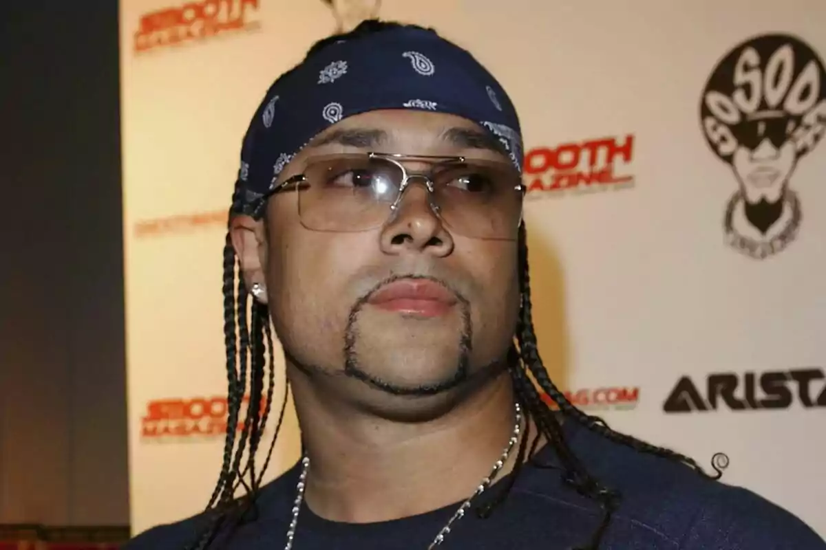 Fotografía de Chino XL con gafas y una bandana azul con diseños blancos, con trenzas en el cabello y una barba delineada, posando frente a un fondo con logotipos de Smooth Magazine y Arista.