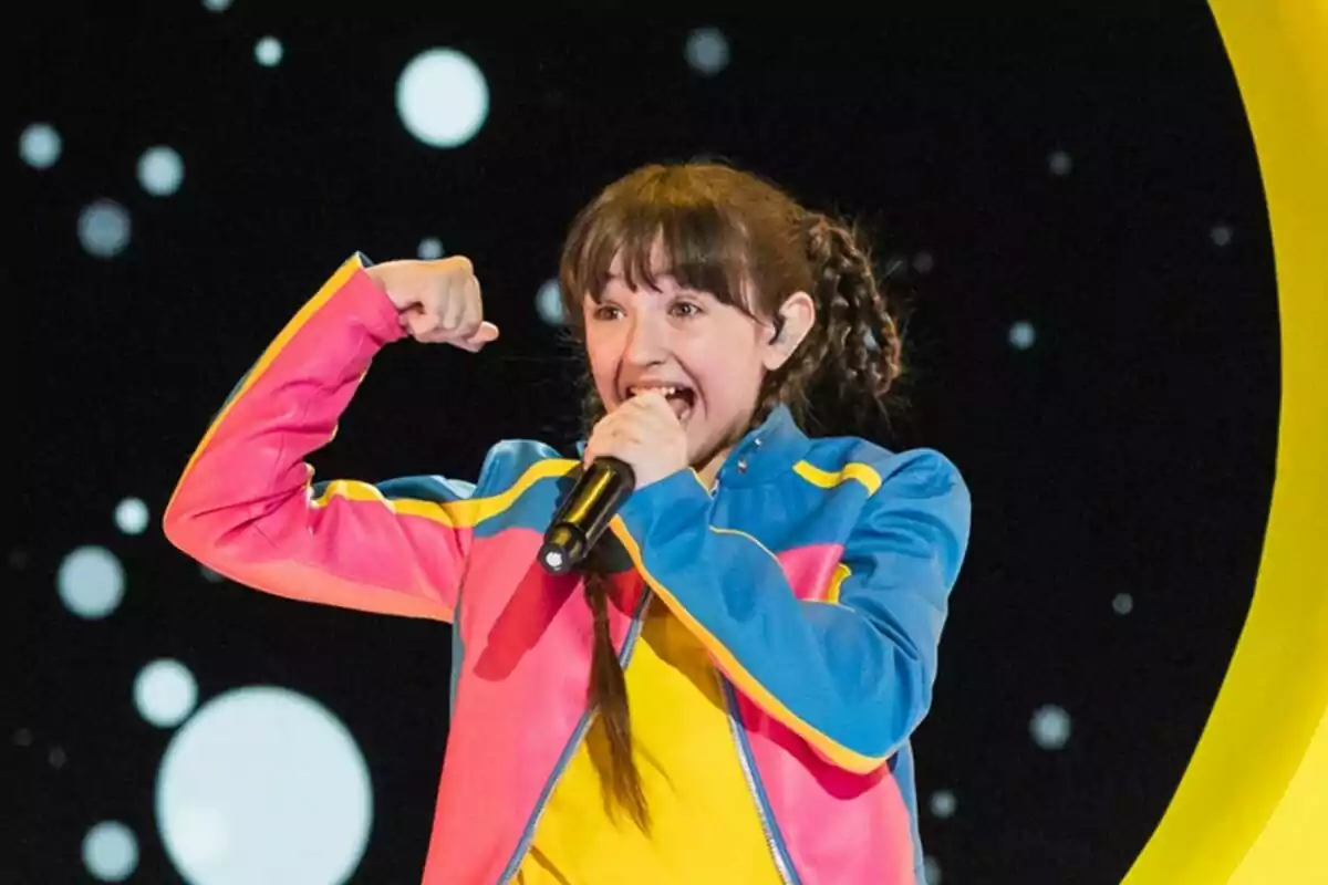 Chloe Delarosa con chaqueta colorida canta emocionada en un escenario con fondo de círculos blancos en Eurovisión Junior.