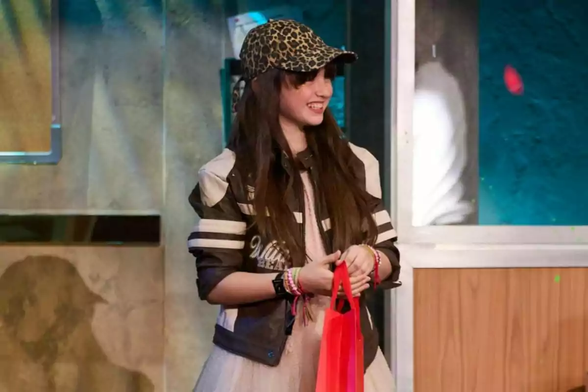 Chloe DelaRosa con gorra de estampado animal y chaqueta a rayas sostiene una bolsa roja mientras sonríe en un entorno interior en La Revuelta.