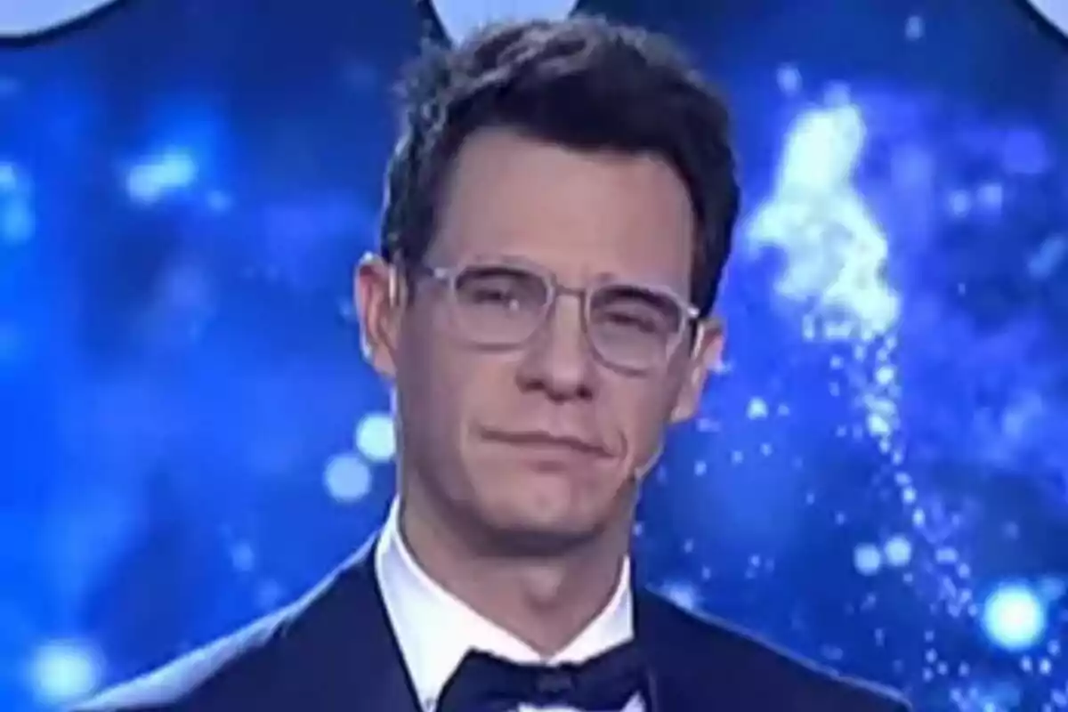 Christian Gálvez con gafas y esmoquin frente a un fondo azul con luces brillantes en el especial de Nochebuena de Telecinco.