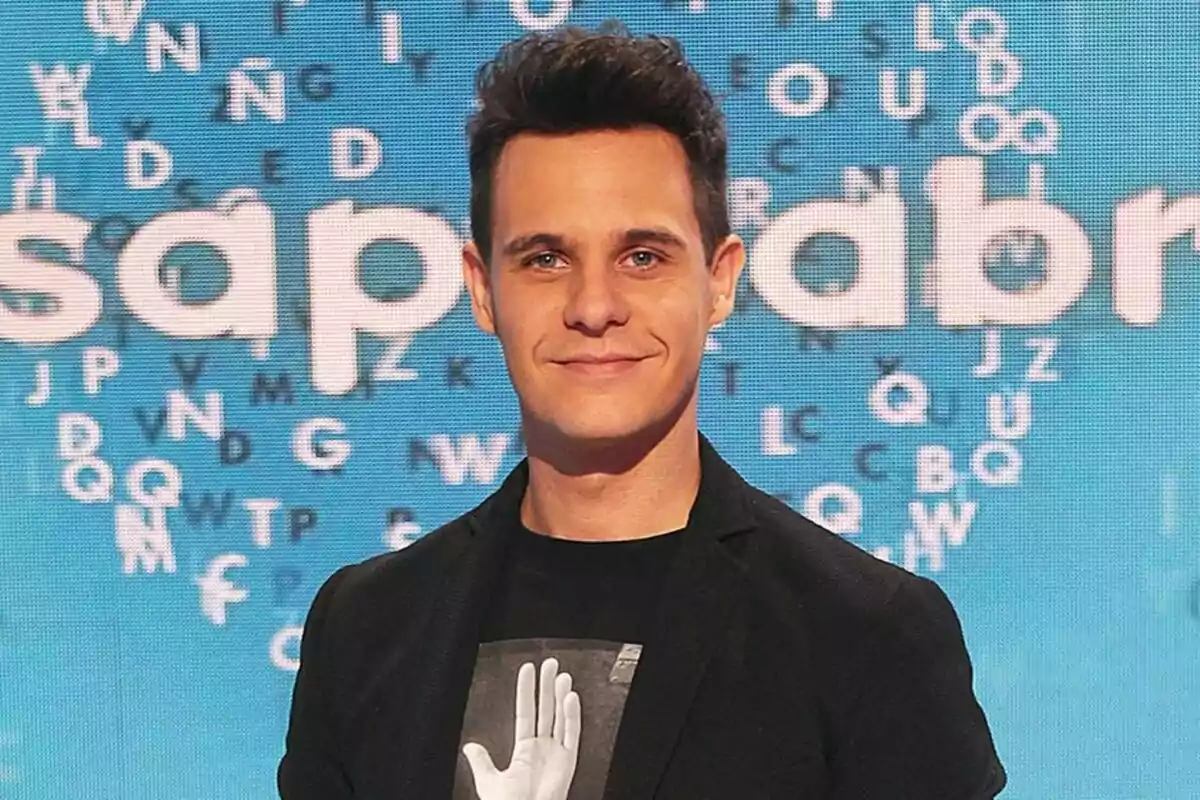 Christian Gálvez con cabello oscuro y una chaqueta negra está de pie frente a un fondo azul con letras dispersas en Pasapalabra.