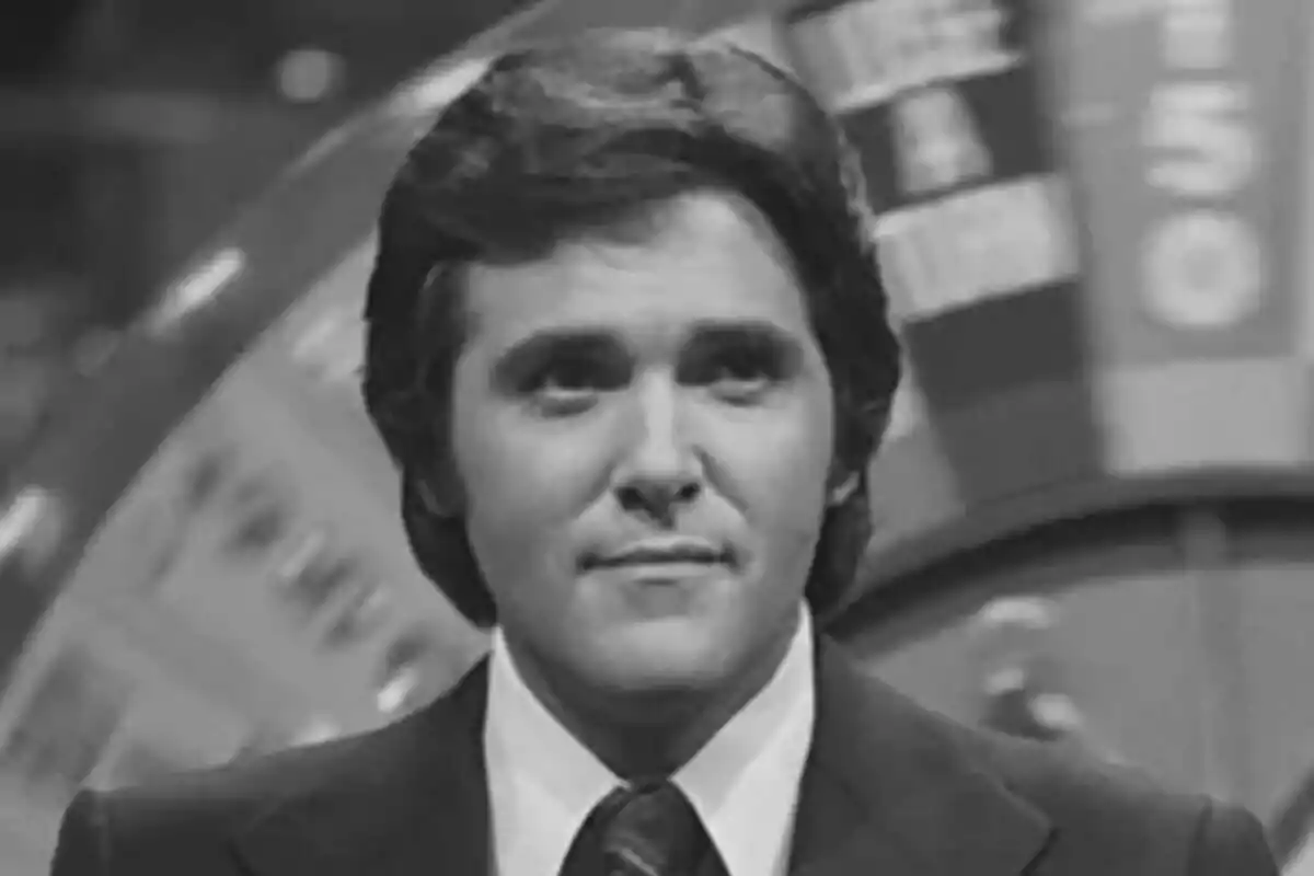 Chuck Woolery con traje y corbata frente a una rueda de la fortuna en blanco y negro.