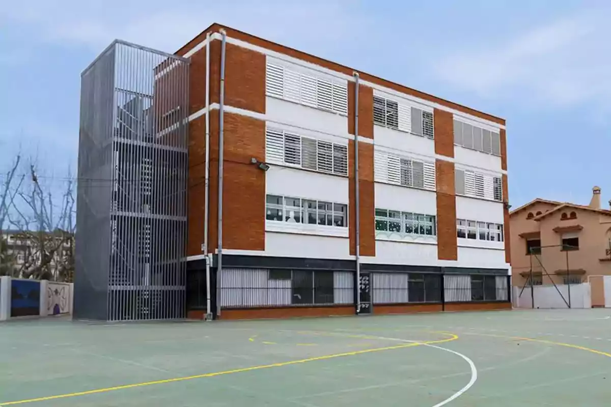 Edificio escolar de ladrillo rojo con ventanas blancas y una estructura metálica en el lateral, situado junto a una cancha deportiva. Edificio escolar de ladrillo rojo con ventanas blancas y una estructura metálica en el lateral, situado junto a una cancha deportiva.