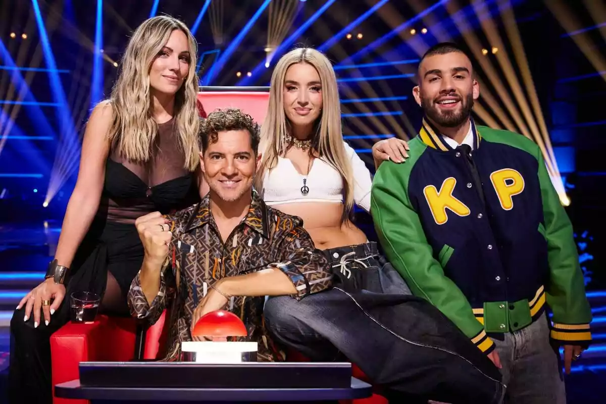 Edurne, David Bisbal, Lola Índigo y Manuel Turizo posan juntas en un escenario iluminado con luces azules y amarillas, una de ellas está sentada en una silla roja con un botón grande frente a ellos en La Voz Kids. Edurne, David Bisbal, Lola Índigo y Manuel Turizo posan juntas en un escenario iluminado con luces azules y amarillas, una de ellas está sentada en una silla roja con un botón grande frente a ellos en La Voz Kids.