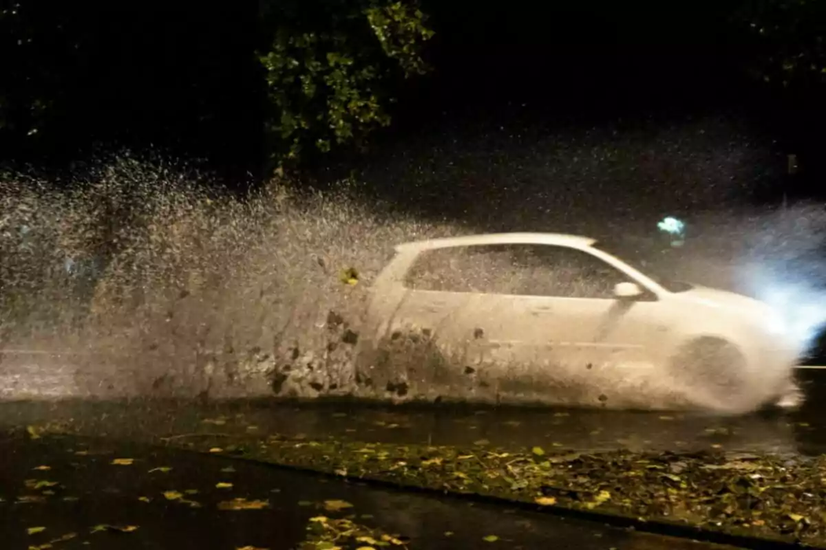 Un coche blanco salpicando agua mientras pasa por un charco en una calle oscura. Un coche blanco salpicando agua mientras pasa por un charco en una calle oscura.