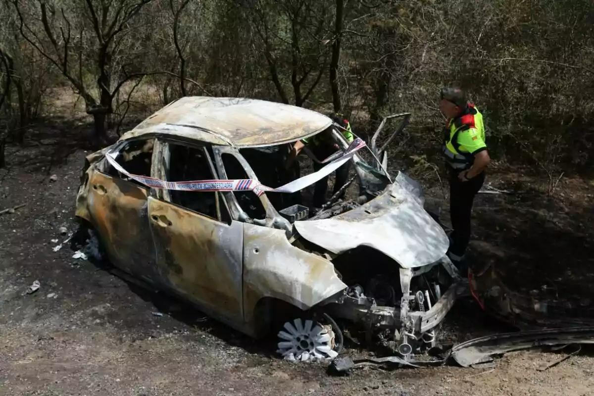 Un coche quemado y destrozado en un área boscosa, con dos personas de pie cerca del vehículo inspeccionándolo. Un coche quemado y destrozado en un área boscosa, con dos personas de pie cerca del vehículo inspeccionándolo.