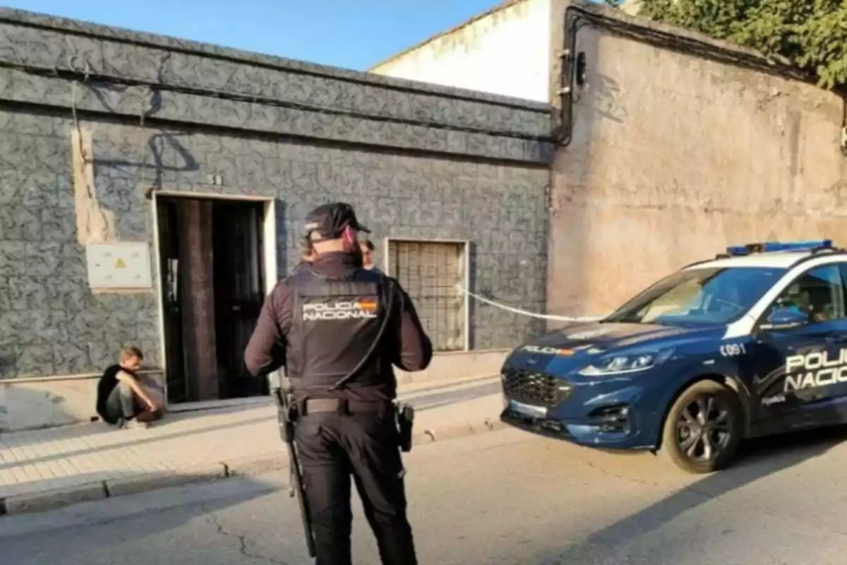 Un agente de la Policía Nacional está de pie frente a un edificio, mientras un coche patrulla está estacionado cerca y una persona está sentada en la acera.