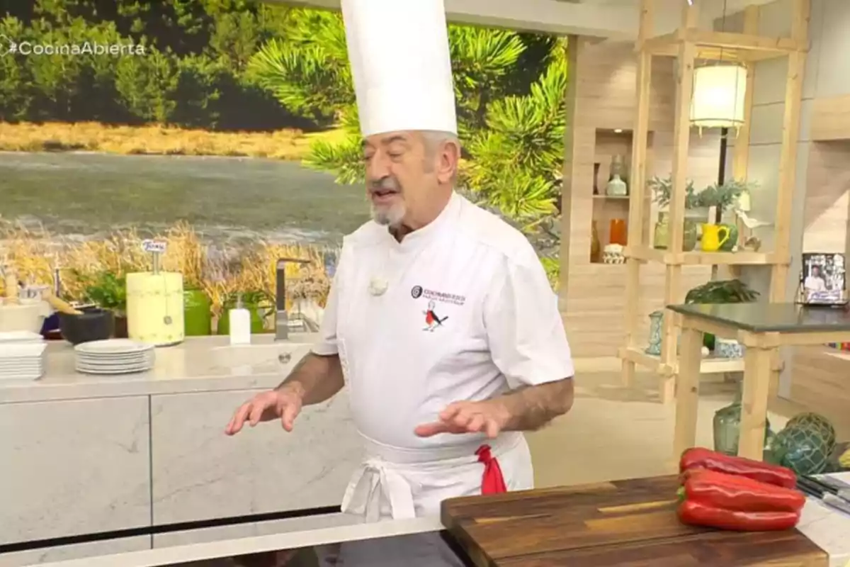 Karlos Arguiñano con uniforme blanco y gorro alto está en una cocina moderna con un fondo de paisaje natural, junto a una tabla de cortar con pimientos rojos. Captura de 'Cocina Abierta' Karlos Arguiñano con uniforme blanco y gorro alto está en una cocina moderna con un fondo de paisaje natural, junto a una tabla de cortar con pimientos rojos. Captura de 'Cocina Abierta'
