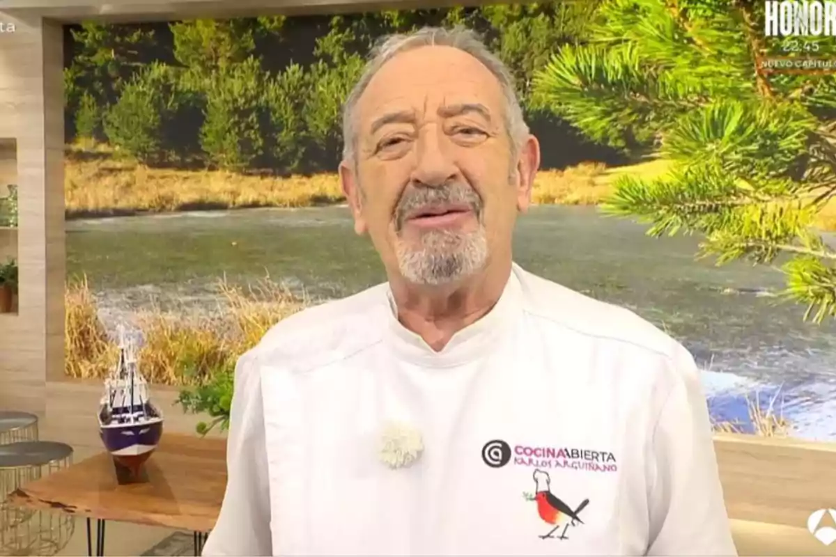 Karlos Arguiñano con barba y bigote está de pie en un estudio de televisión con un fondo de paisaje natural y lleva una chaqueta de chef blanca con un logotipo bordado. Captura de 'Cocina Abierta'
