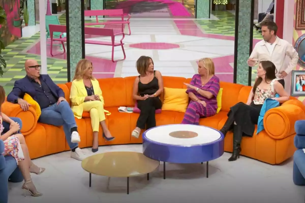 Un grupo de personas formado por Kiko Matamoros, Lydia Lozano, María Patiño, Belén Esteban, Inés Hernand y Aitor Albizua está sentado en un sofá naranja en un set de televisión de La Familia de la Tele colorido, conversando entre ellos mientras un hombre de pie parece estar hablando.