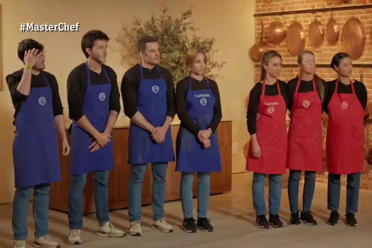 Seis personas con delantales de color azul y rojo están formadas en dos equipos dentro de una cocina con decoración de cobre y el logo de MasterChef aparece en la esquina superior izquierda Seis personas con delantales de color azul y rojo están formadas en dos equipos dentro de una cocina con decoración de cobre y el logo de MasterChef aparece en la esquina superior izquierda
