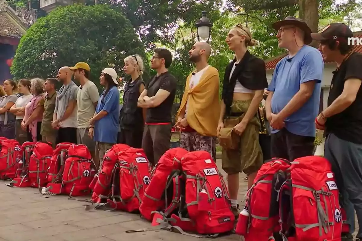 Un grupo de concursantes de pie en fila junto a mochilas rojas en un entorno al aire libre con árboles y un edificio al fondo en la promo de Pekin Express.