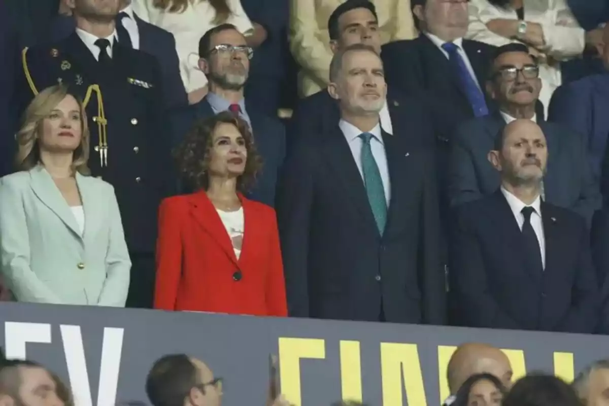 Un grupo de personas vestidas formalmente está de pie en un evento, observando atentamente algo frente a ellos. Un grupo de personas vestidas formalmente está de pie en un evento, observando atentamente algo frente a ellos.