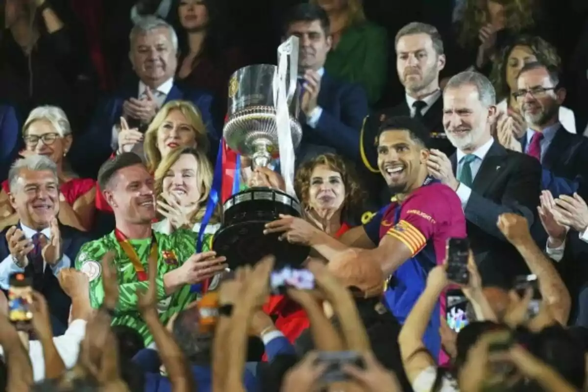 Un grupo de personas celebra mientras dos jugadores sostienen un trofeo grande y plateado, rodeados de aplausos y sonrisas.