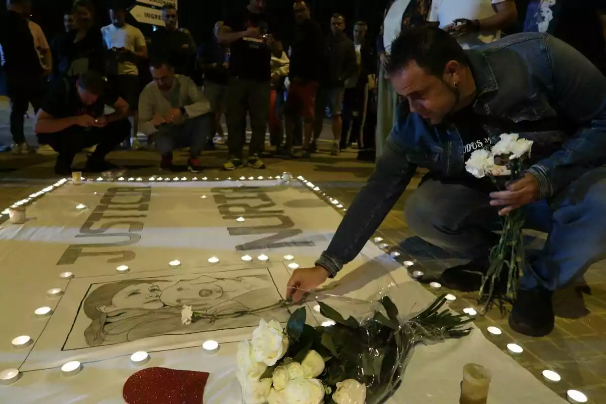 Un grupo de personas se reúne en una vigilia nocturna, encendiendo velas y colocando flores alrededor de un cartel que pide justicia.