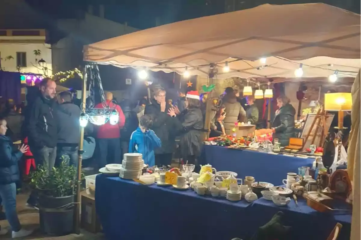 Una feria nocturna al aire libre con personas comprando en un puesto iluminado que vende artículos diversos sobre una mesa cubierta con un mantel azul. Una feria nocturna al aire libre con personas comprando en un puesto iluminado que vende artículos diversos sobre una mesa cubierta con un mantel azul.