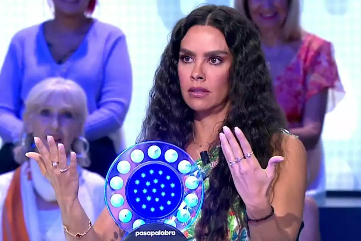 Cristina Pedroche con cabello rizado y expresión seria sostiene un objeto circular iluminado con luces azules en un programa de televisión llamado "Pasapalabra" mientras otras personas observan en el fondo.