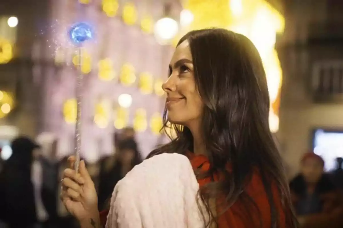 Cristina Pedroche sonriente sostiene una varita mágica con una luz azul brillante en un entorno urbano iluminado.