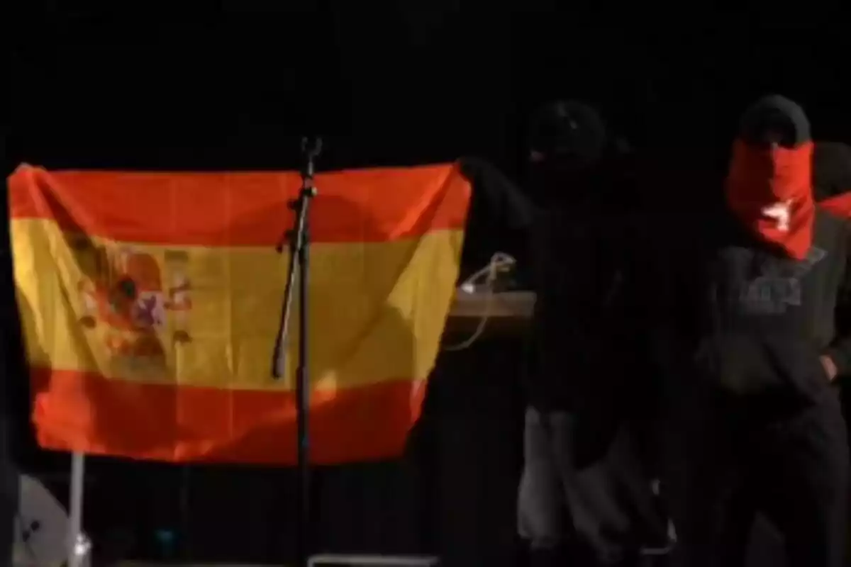 Personas enmascaradas sosteniendo una bandera de España junto a un micrófono en un escenario oscuro.