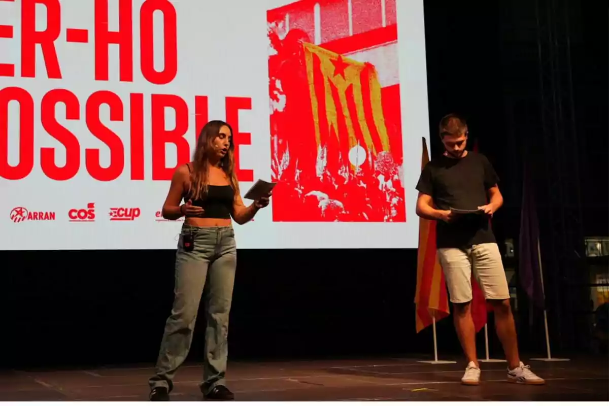 Dos personas en un escenario con una pantalla de fondo que muestra un mensaje en catalán y varias banderas.