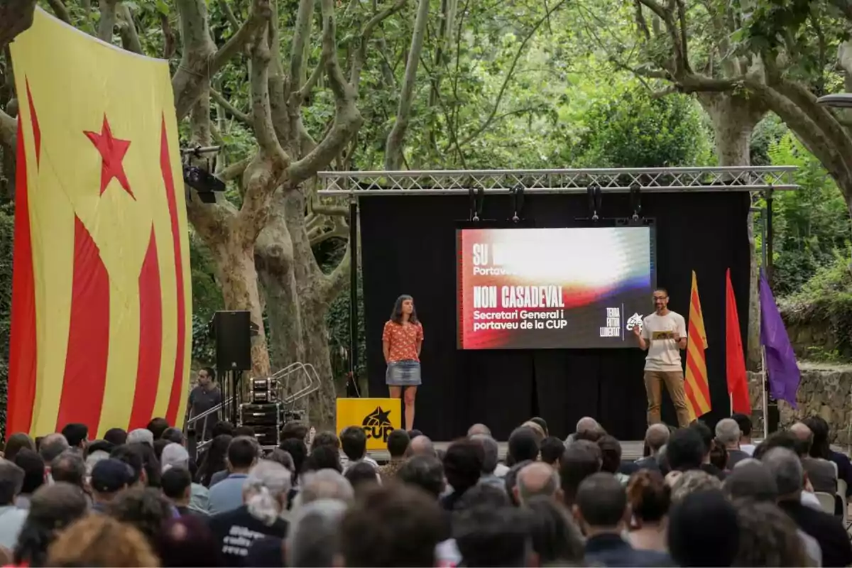 Acto político al aire libre con una gran bandera estelada amarilla, dos personas en un escenario frente a una pantalla y un público numeroso rodeado de árboles Acto político al aire libre con una gran bandera estelada amarilla, dos personas en un escenario frente a una pantalla y un público numeroso rodeado de árboles