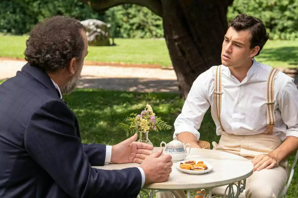 Curro y Alonso conversan en una mesa al aire libre en un jardín, uno de ellos lleva tirantes y una camisa blanca mientras el otro viste un traje oscuro, en la serie La Promesa.