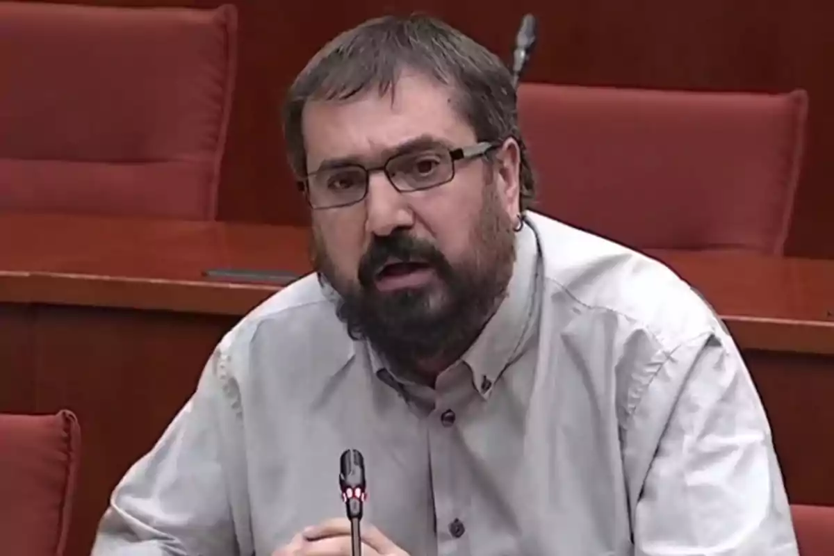 Hombre con barba y gafas hablando en un micrófono en una sala de conferencias.