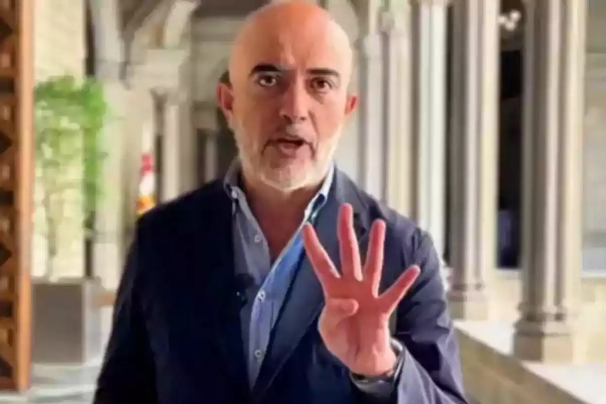 Hombre calvo con barba y chaqueta azul levantando cuatro dedos en un edificio con columnas al fondo