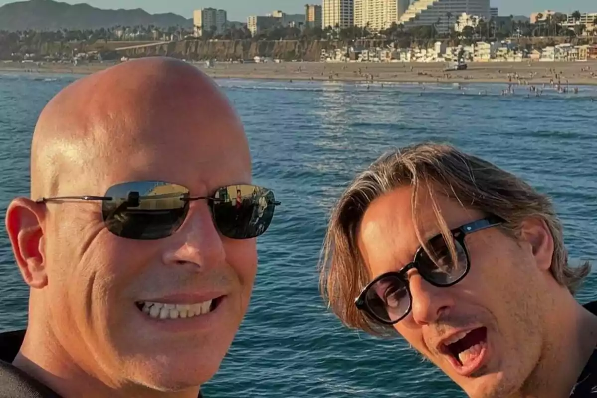 Alberto Caballero junto a Daniel Deorador con gafas de sol posan para una selfie frente al mar con una ciudad y montañas al fondo.