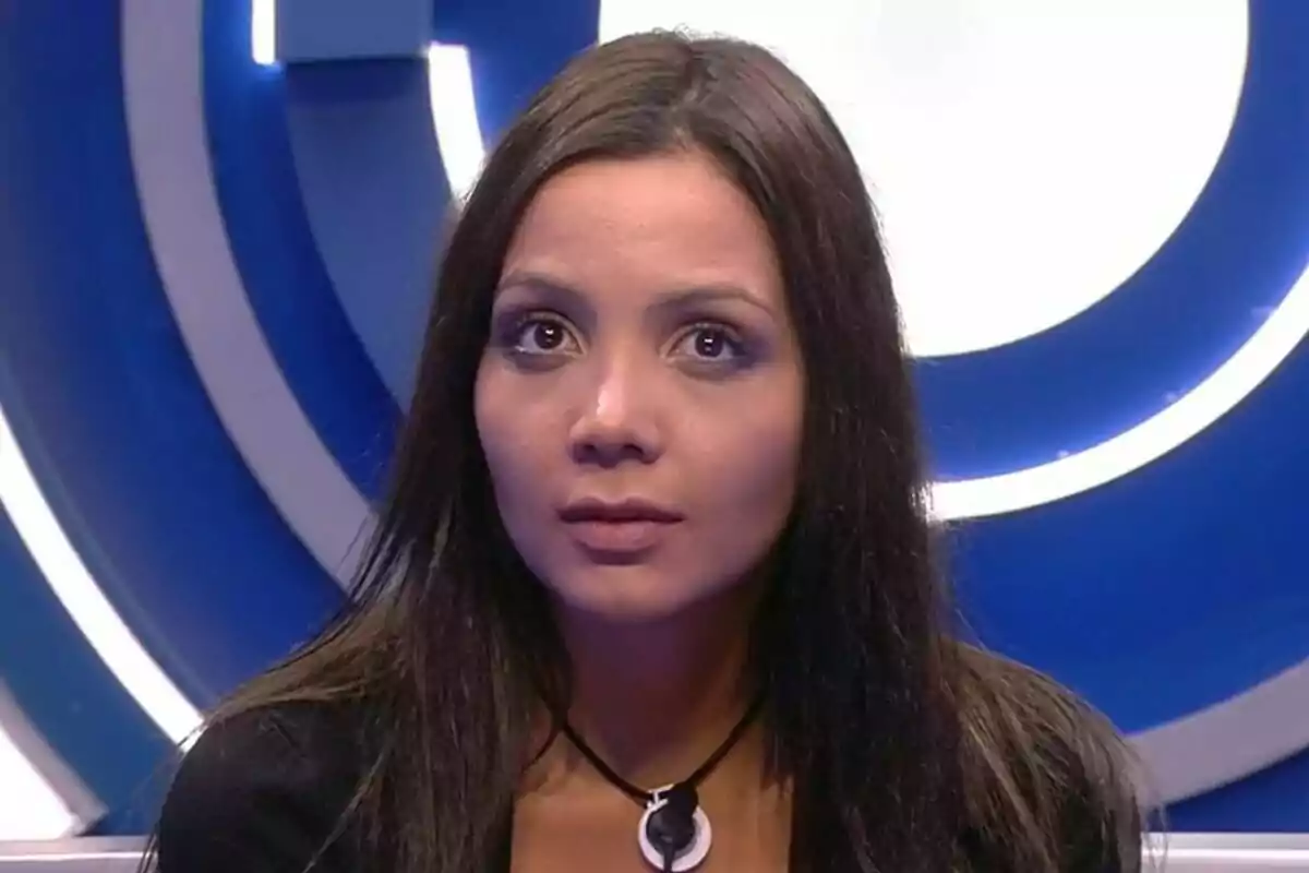 Daniela con cabello oscuro y expresión seria frente a un fondo azul con un diseño circular iluminado en Gran Hermano.