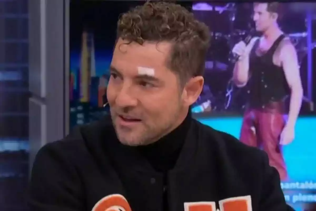 David Bisbal con cabello rizado y una curita en la ceja está hablando en El Hormiguero con una imagen de fondo de una persona cantando.