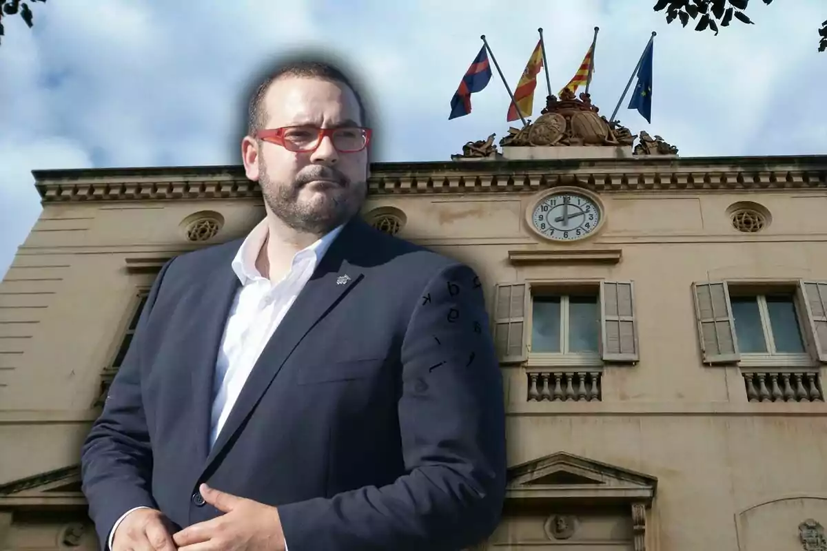 Hombre con gafas rojas y barba frente a un edificio con un reloj y varias banderas ondeando en la parte superior.