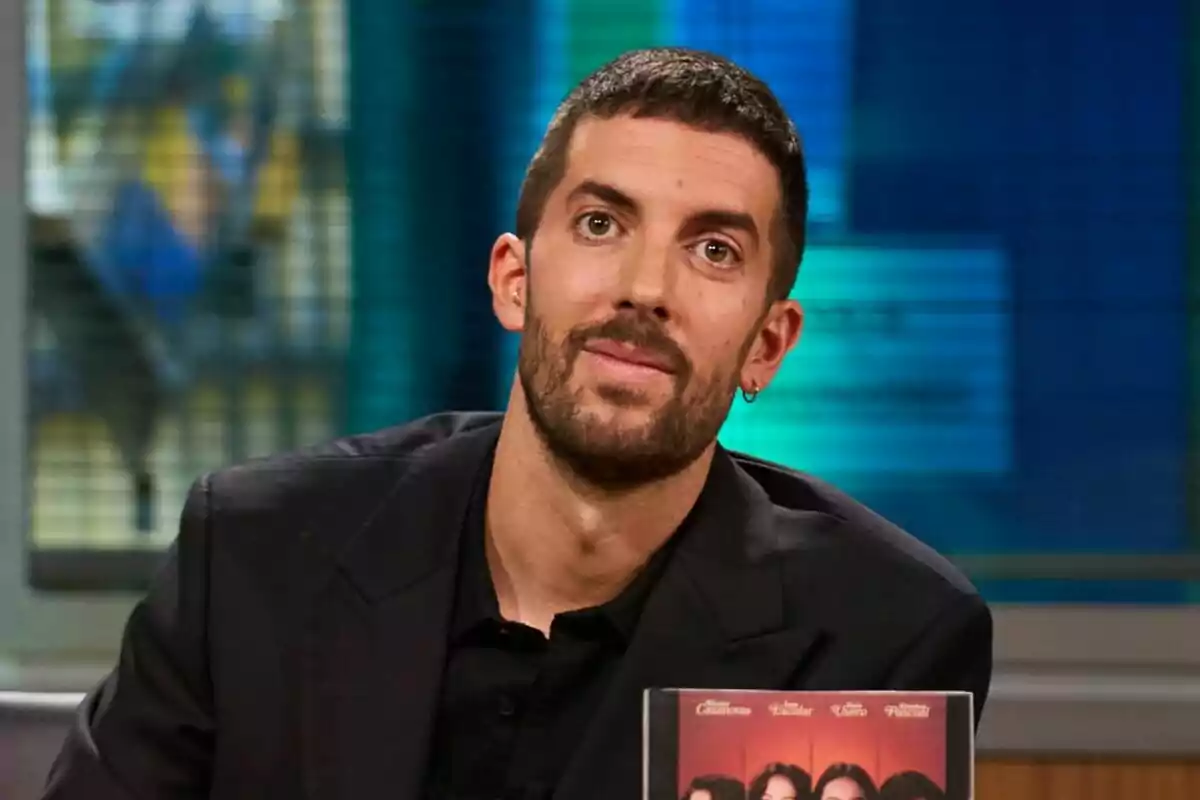 David Broncano con barba y cabello corto, vestido con un traje oscuro está sentado en el plató de La Revuelta con un fondo desenfocado de colores azul y verde.