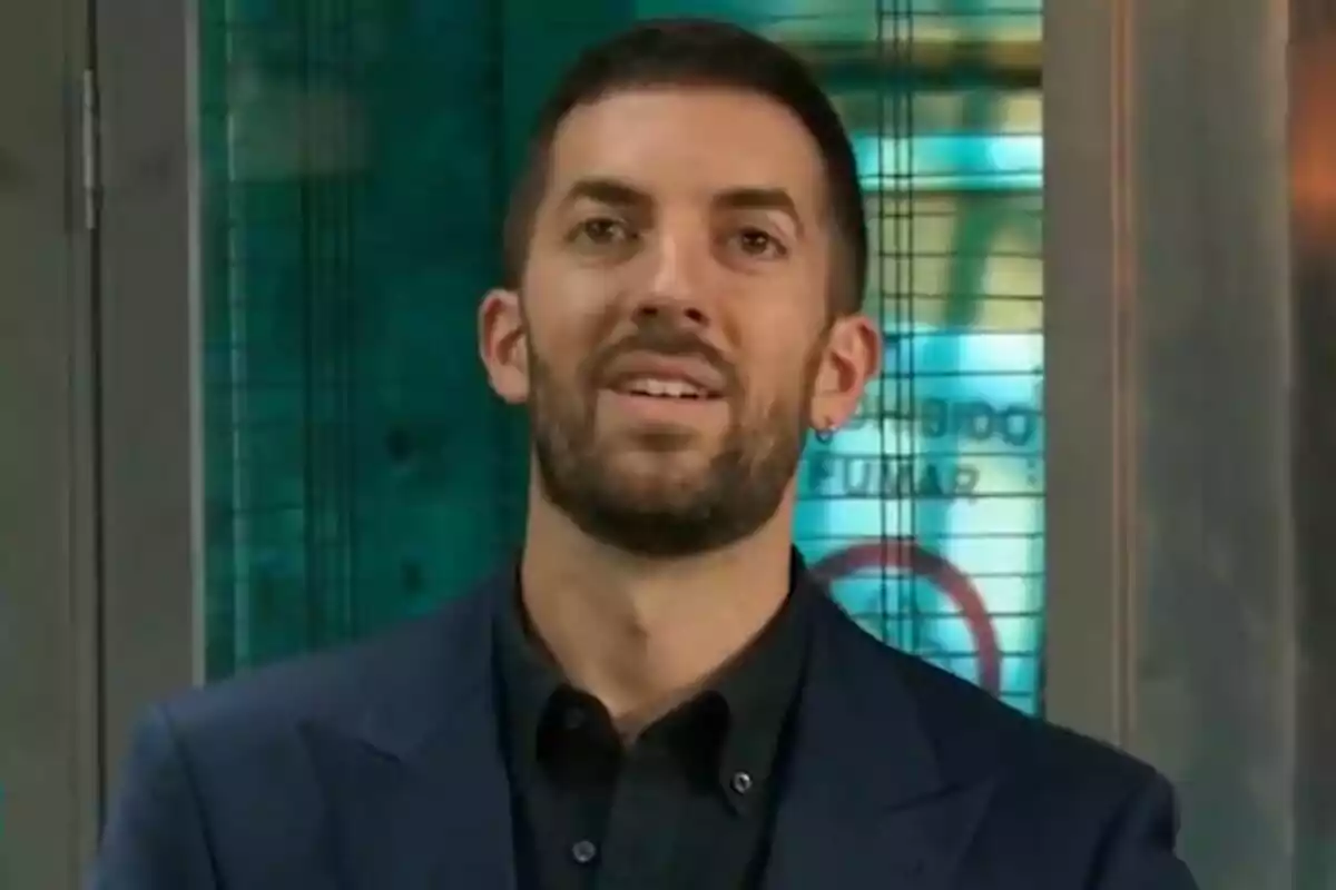 David Broncano de barba corta y cabello oscuro viste saco azul marino y camisa negra frente a un fondo con tonos verdes y azules en La Revuelta