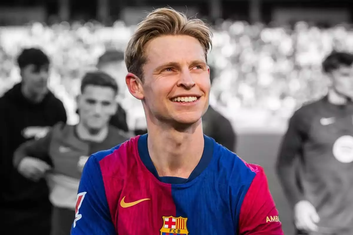 Un jugador de fútbol sonríe mientras lleva una camiseta azul y roja en un campo con personas desenfocadas al fondo.