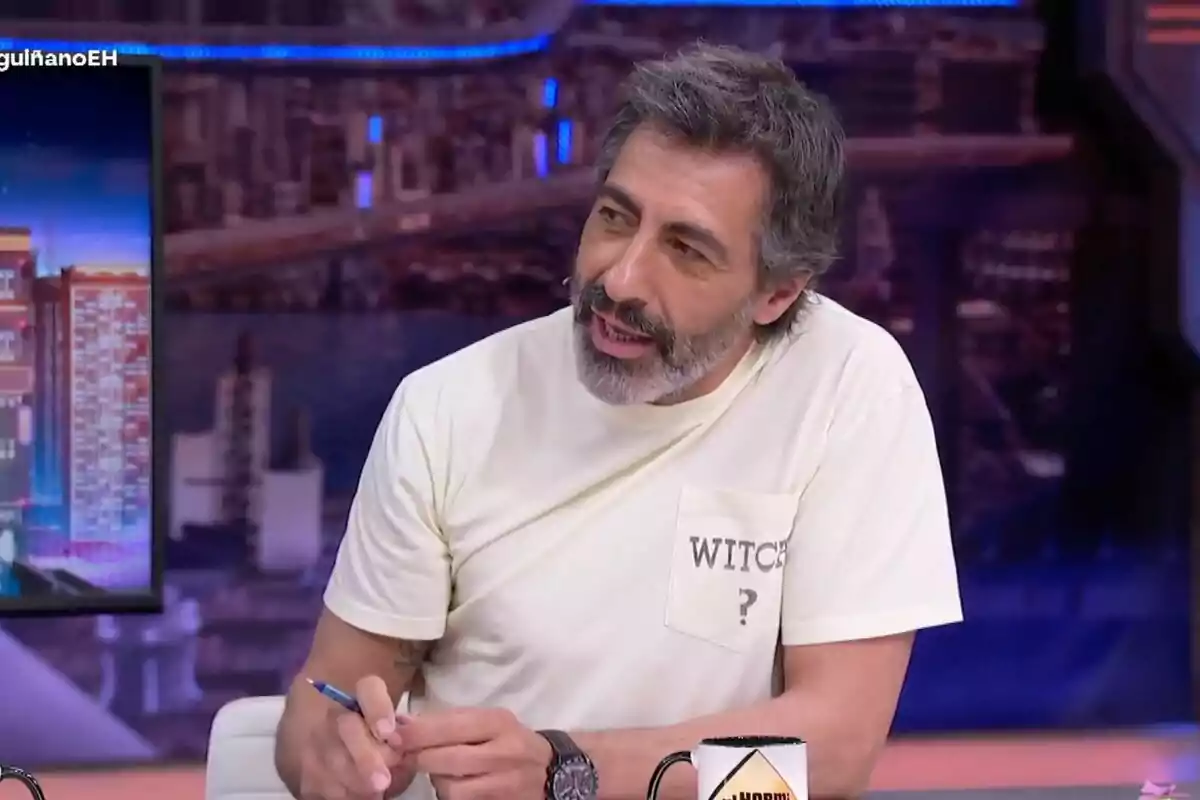 Juan del Val con cabello y barba canosos sentado en 'El Hormiguero', lleva puesta una camiseta clara con la palabra WITCH y un signo de interrogación en el bolsillo, sostiene un bolígrafo y una taza está sobre la mesa frente a él.
