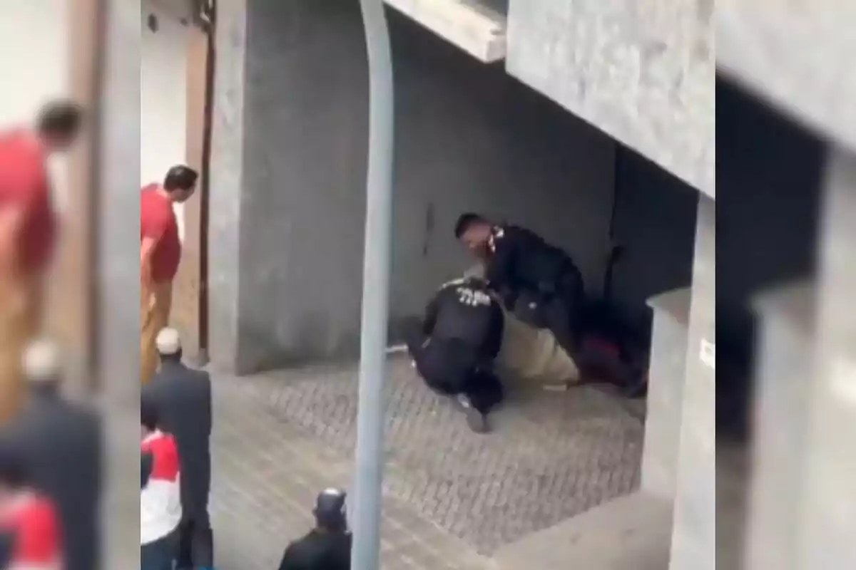 Varias personas observan mientras dos policías detienen a un individuo en la entrada de un edificio.