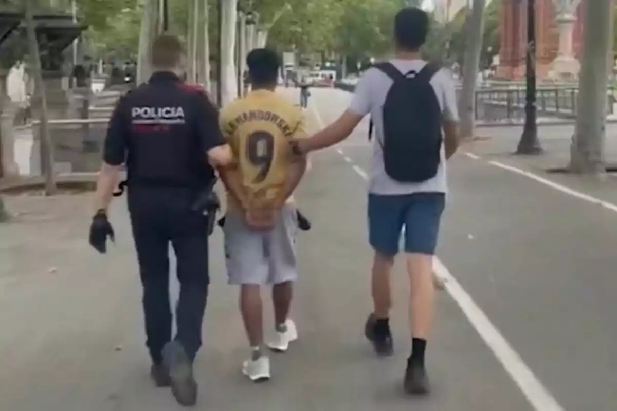 Un policía y otra persona escoltan a un hombre con una camiseta de fútbol mientras caminan por una calle arbolada.