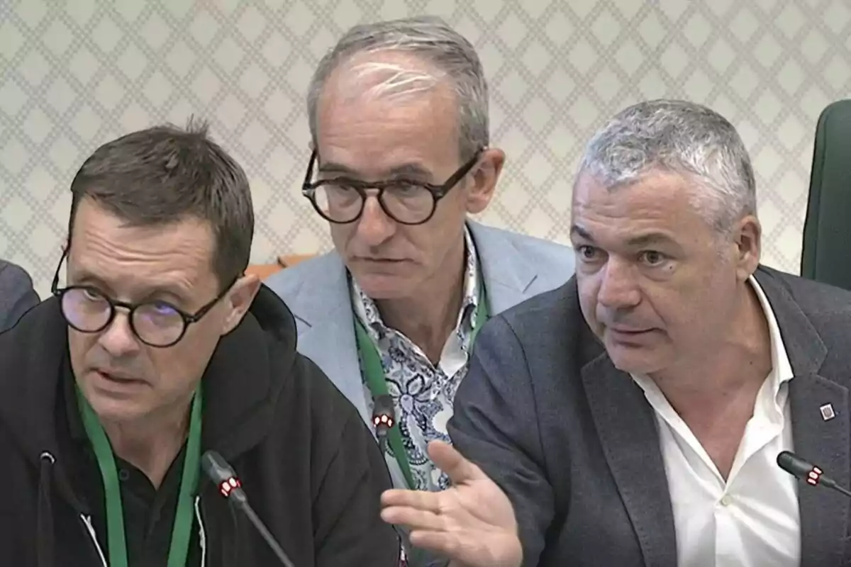 Tres hombres sentados en una mesa de reunión con micrófonos y credenciales verdes, conversando de manera seria.